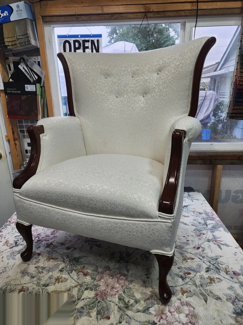 Antique wingback chair reupholstered in cream jacquard fabric, staged with the shop sign.